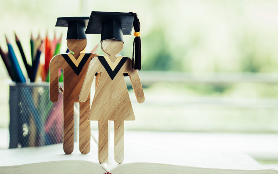 Two wooden people figurines with graduation caps on a desk top.